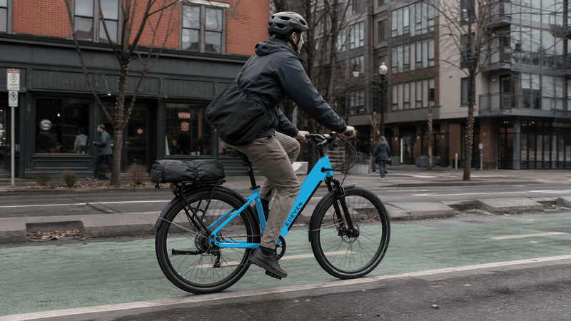 E11EVEN DRAGONFLY electric bike in bright blue with step-thru aluminum frame, rear cargo rack, and black tires ridden by a cyclist.
