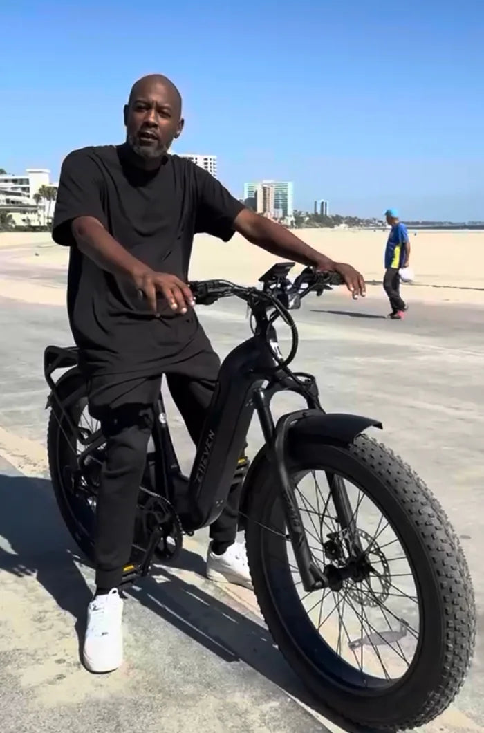 Rider in black with a black E11EVEN fat-tire electric bike on a beach promenade.