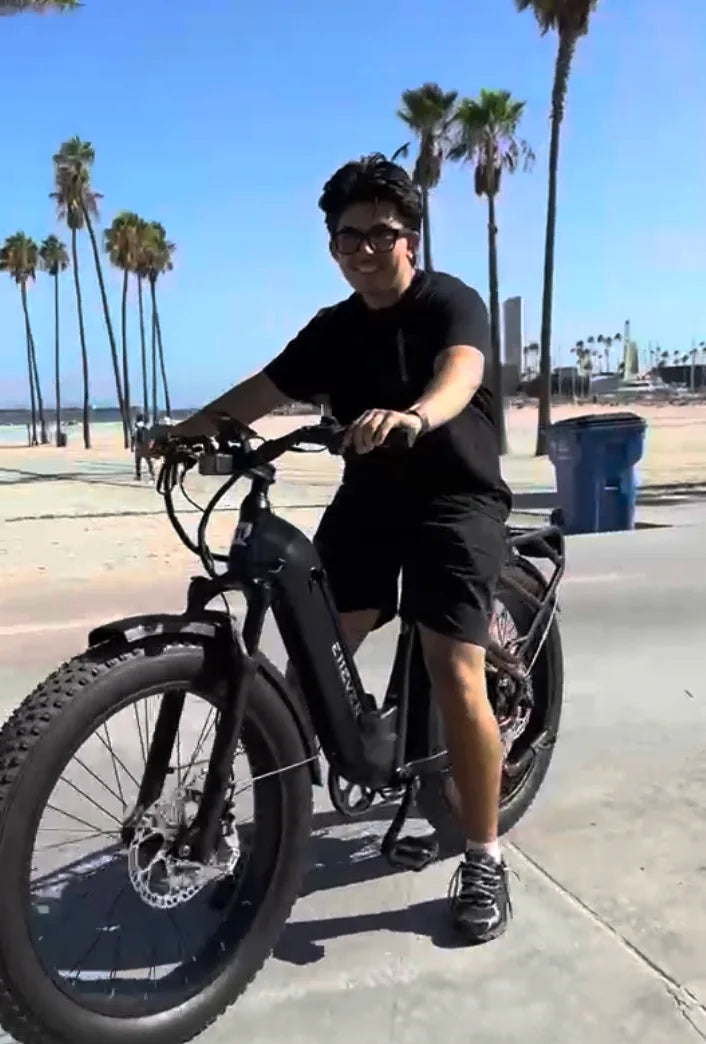 Rider wearing sunglasses on a black E11EVEN fat-tire electric bike beside a palm-lined beach.
