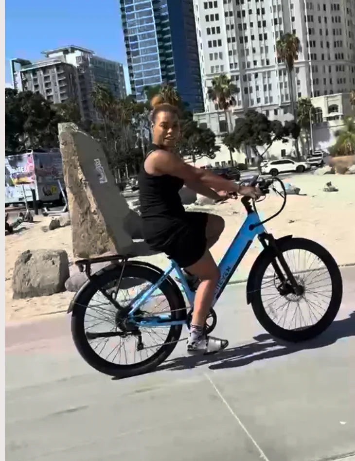 Rider on a blue E11EVEN step-through electric bike near beachfront buildings.