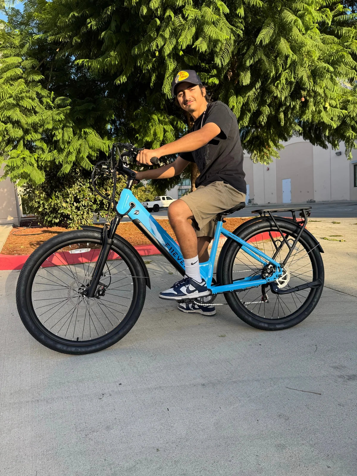 Rider on a blue E11EVEN step-through electric bike beside a tree-lined street.