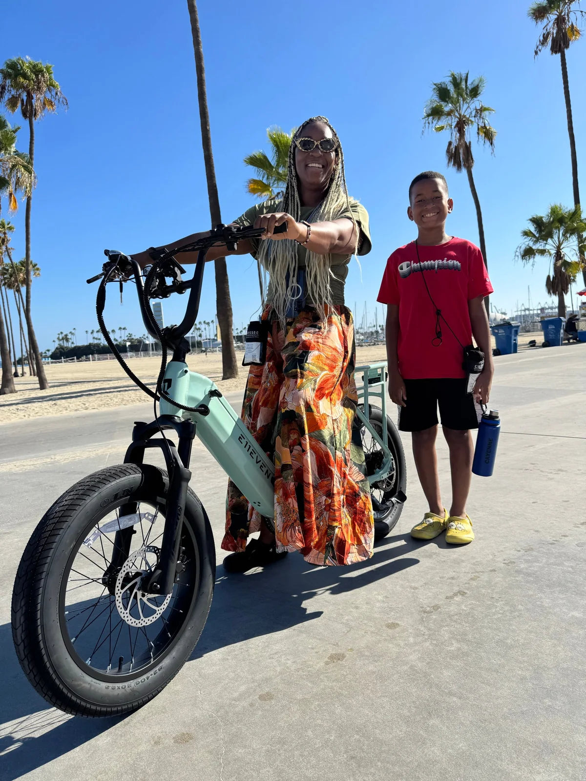 Two people posing with a mint-green E11EVEN fat-tire electric bike beside a palm-lined beach.