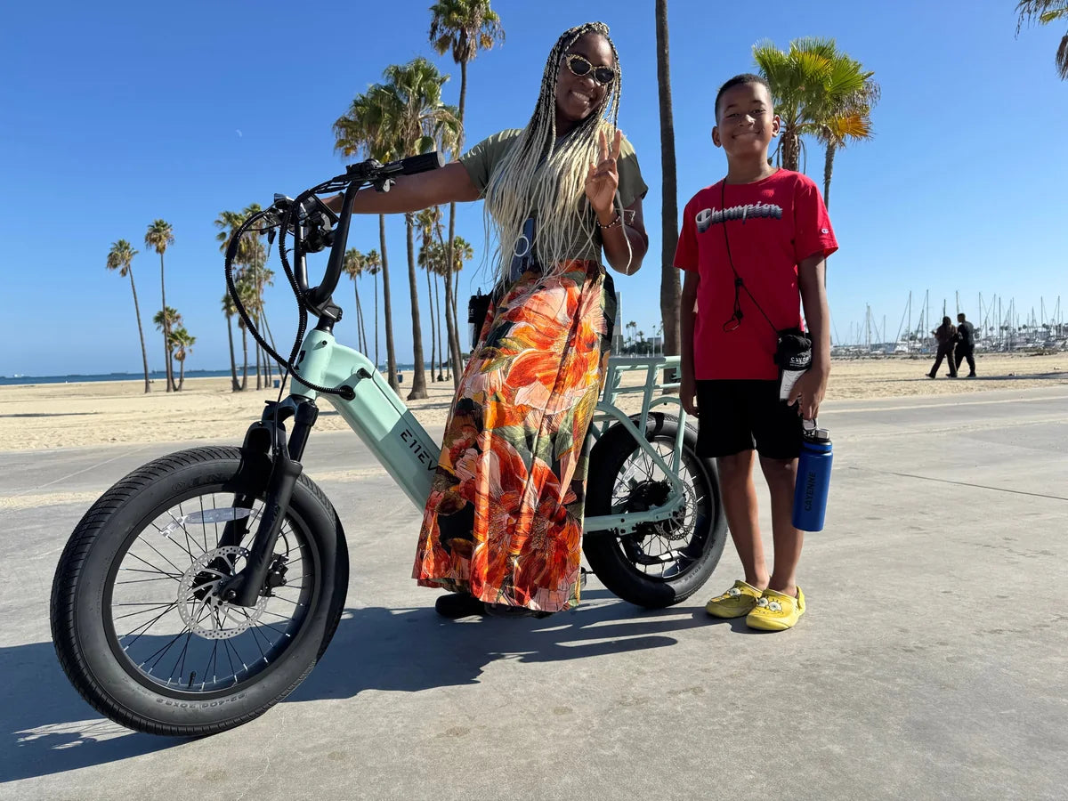 Two people beside a mint-green E11EVEN fat-tire electric bike on a palm-lined beachfront.