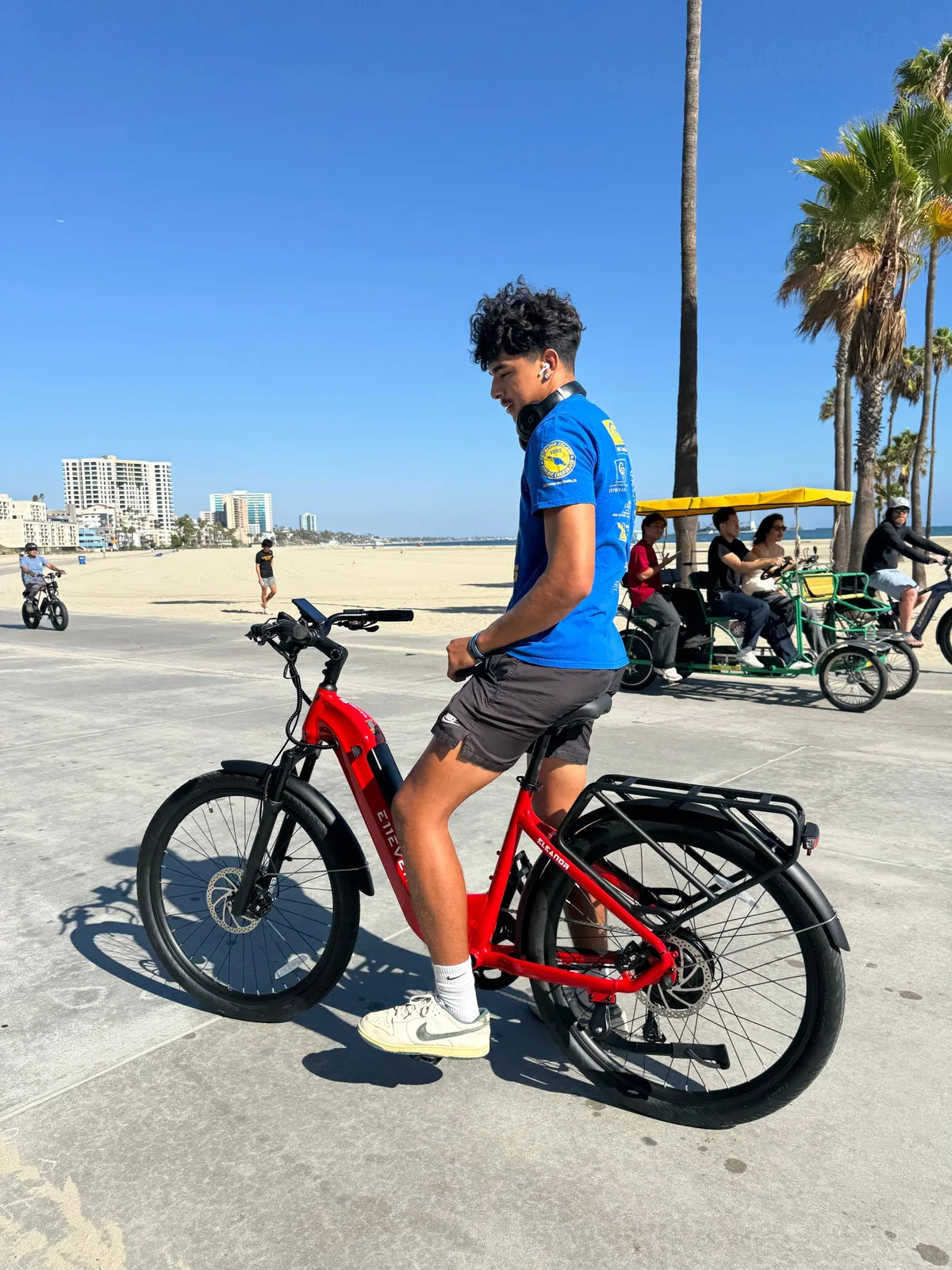 Rider in a blue shirt seated on a red E11EVEN step-through electric bike by the beach.