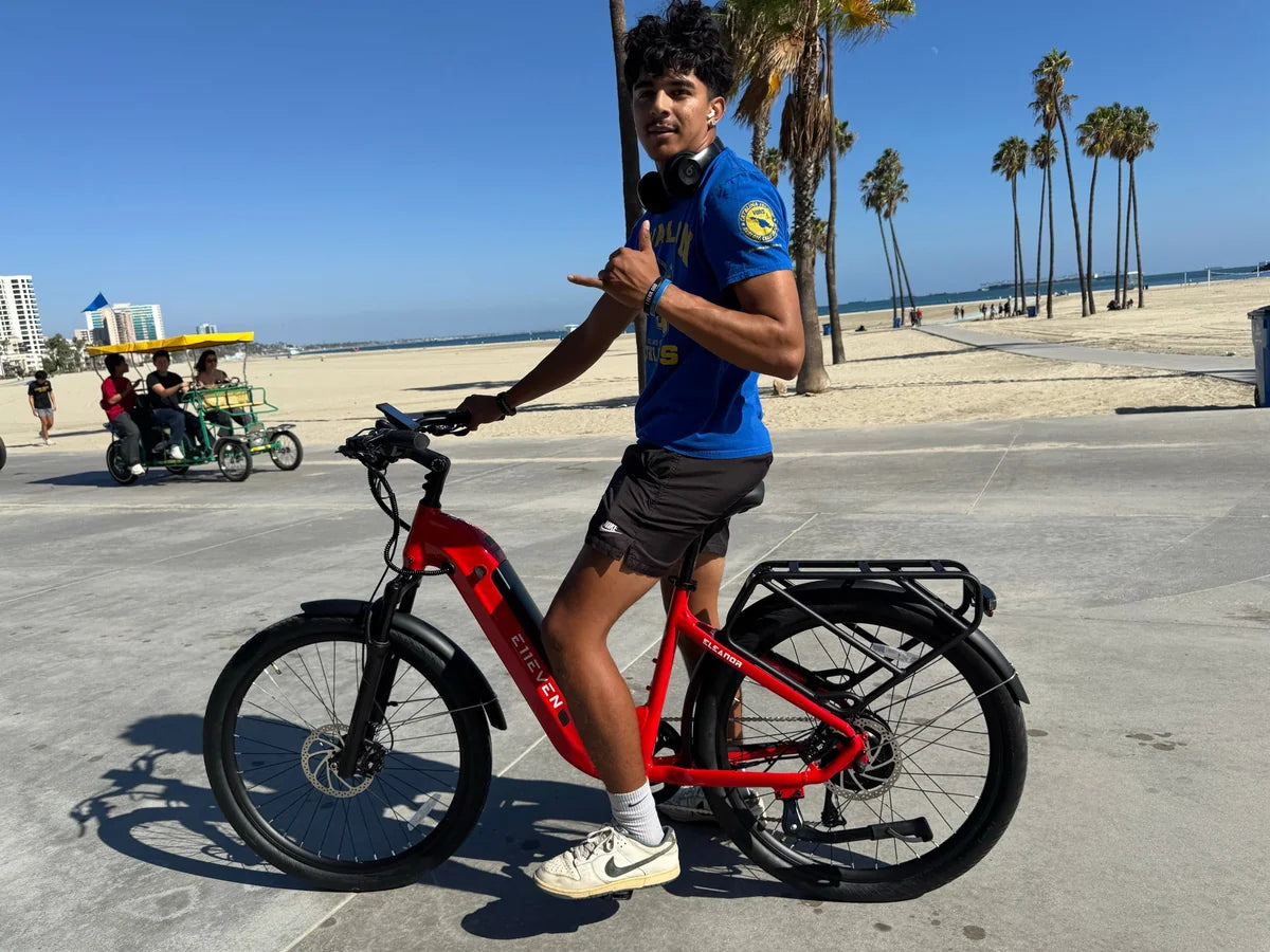 Rider in a blue shirt with a red E11EVEN step-through electric bike on a beachfront promenade.
