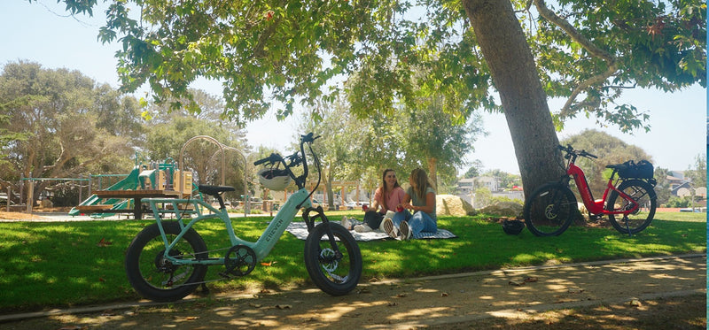Two women sitting on a blanket in a park beside the E11EVEN WINGMAN and the E11EVEN ELEANOR electric bike